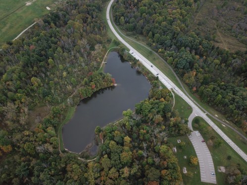 Aerial view of a winding road beside a serene pond surrounded by lush trees in autumn colors.