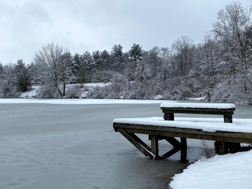 A snowy landscape featuring a frozen lake and a wooden dock, surrounded by trees and a cloudy sky.