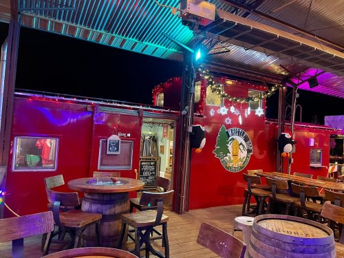 A vibrant outdoor bar with colorful lights, wooden tables, and a red shipping container backdrop.