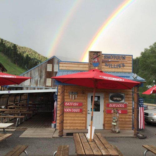 A rustic wooden restaurant with red umbrellas, featuring a rainbow in the background and a sign for burgers and catfish.