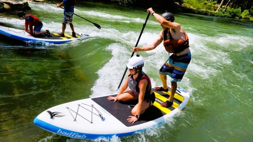 A person paddles on a stand-up paddleboard while another sits on the board, navigating a river with gentle waves.