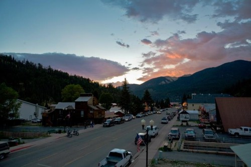 A scenic view of a small town street at sunset, surrounded by mountains and trees, with parked cars and buildings.