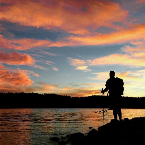 A silhouetted hiker stands by a lake at sunset, with vibrant orange and pink clouds reflecting in the water.