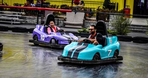 Two people race in colorful go-karts on a track, one in a purple car and the other in a teal car.