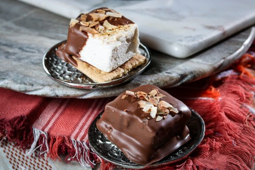 Two chocolate-covered marshmallow squares on decorative plates, garnished with almond slivers, on a textured surface.