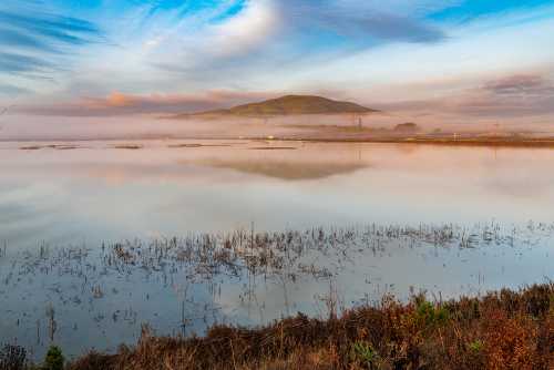 A serene landscape featuring a misty lake, distant hills, and a colorful sky reflecting on the water's surface.