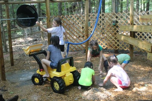 Children playing in a sandy outdoor area, using toys and water, with a wooden structure in the background.