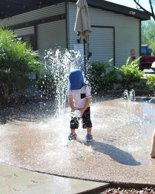 A child in a blue hat plays in a splash pad, surrounded by water jets and greenery on a sunny day.