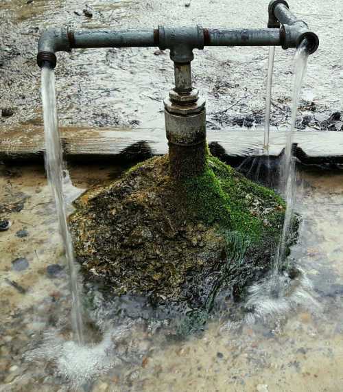 A rusty pipe fountain with water flowing from two spouts, surrounded by moss and rocks.