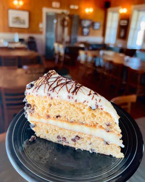 A slice of layered cake with chocolate chips and frosting, served on a black plate in a cozy dining setting.