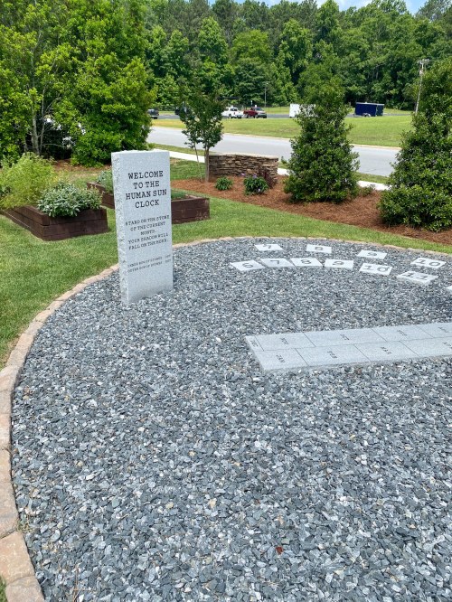 A stone sundial surrounded by gravel and greenery, with a sign reading "Welcome to the Human Sundial."