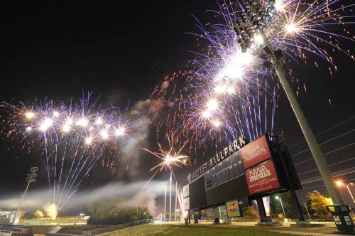 Colorful fireworks illuminate the night sky above a sports stadium, with bright lights and a scoreboard in view.