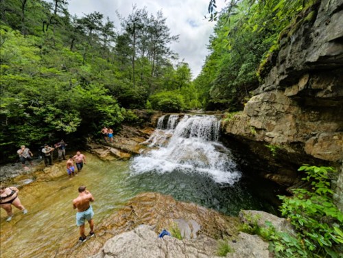 A scenic waterfall surrounded by lush greenery, with people enjoying the water and rocky terrain.