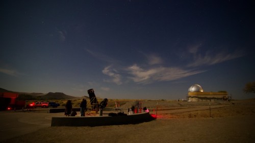 A night scene at an observatory with telescopes, people observing the sky, and a dome structure under a starry sky.