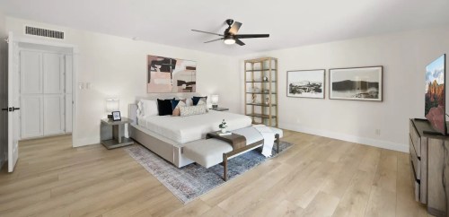 A modern bedroom featuring a large bed, wooden flooring, a ceiling fan, and decorative artwork on the walls.