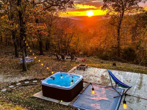 A hot tub on a patio surrounded by trees, with a sunset view and string lights illuminating the scene.
