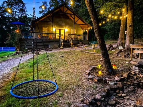 A cozy cabin at dusk, illuminated by string lights, with a swing and garden lights in a serene outdoor setting.
