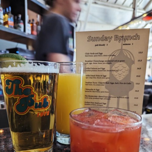Three drinks on a table: a beer, a mimosa, and a cocktail, with a brunch menu in the background.