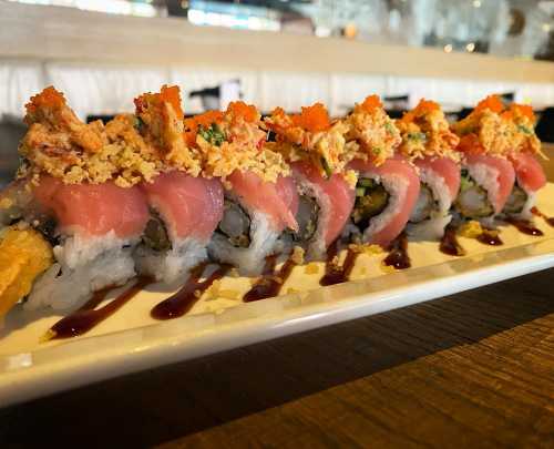 A close-up of a sushi roll topped with crab, fish roe, and crunchy bits, drizzled with sauce on a white plate.