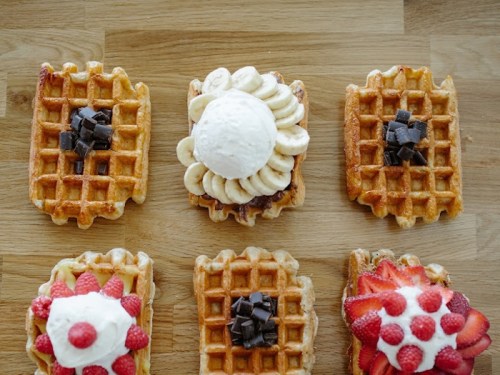 A wooden surface with six waffles topped with ice cream, bananas, chocolate, strawberries, and raspberries.