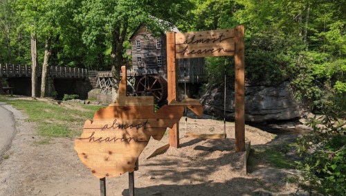Wooden sign shaped like West Virginia with "almost heaven" text, near a rustic building and greenery.