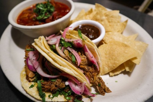 A plate with two tacos filled with meat and onions, served with a bowl of soup and tortilla chips on the side.