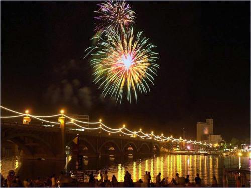 Colorful fireworks illuminate the night sky above a bridge, reflecting on the water with a festive crowd below.