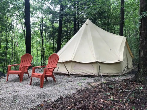 A beige tent in a forested area, with two red Adirondack chairs in front on a gravel surface.