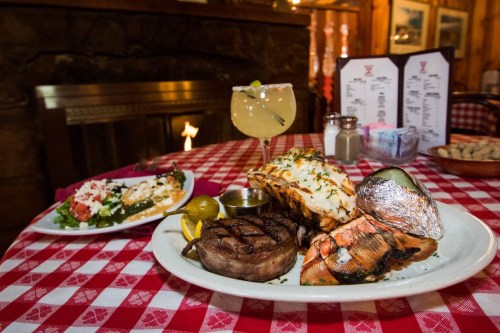 A delicious meal featuring steak, lobster, baked potato, and a cocktail, set on a checkered tablecloth by a fireplace.