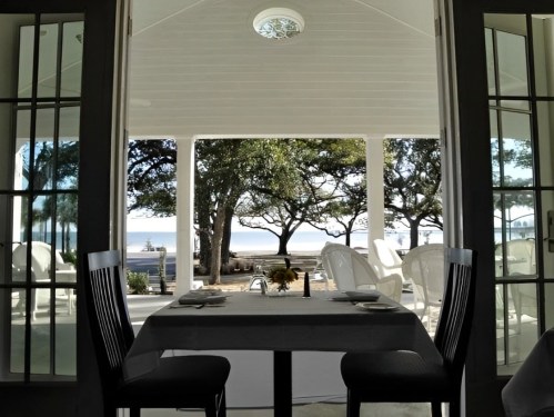 A dining table with two chairs, framed by open doors, overlooking a serene outdoor view with trees and a beach.