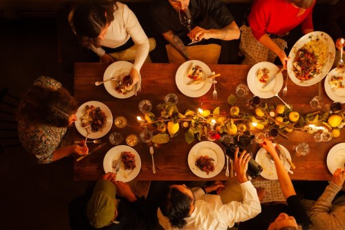Aerial view of a communal dining table with people enjoying a meal, surrounded by candles and decorative foliage.
