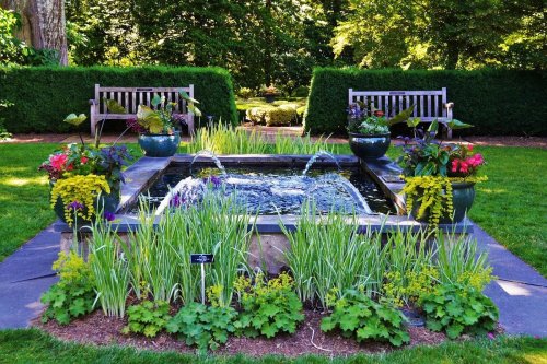 A serene garden scene featuring a pond with fountains, surrounded by vibrant flowers and two wooden benches.