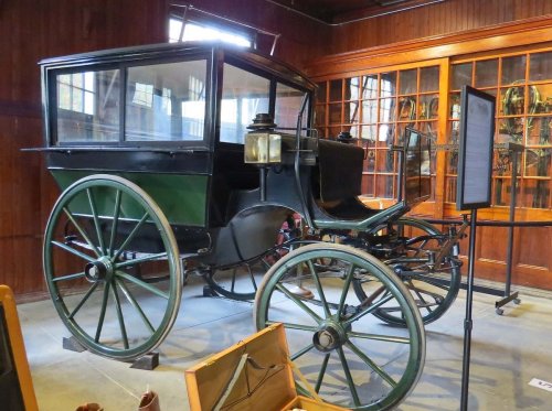 A vintage horse-drawn carriage displayed in a museum, featuring a wooden frame and large wheels.