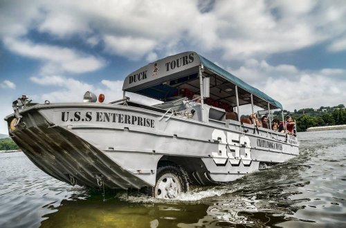 A gray amphibious tour boat named "U.S.S. Enterprise" with passengers, cruising on a calm waterway under a cloudy sky.