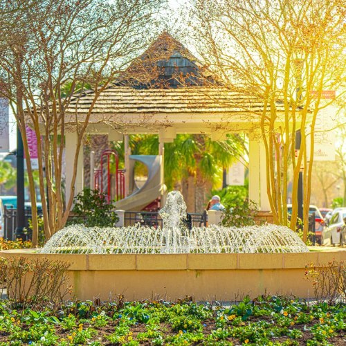 A fountain sprays water in a park, surrounded by trees and a gazebo, with a playground visible in the background.