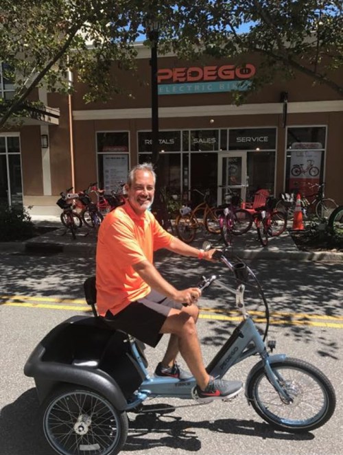 A smiling man in an orange shirt rides an electric tricycle in front of a bike rental shop.