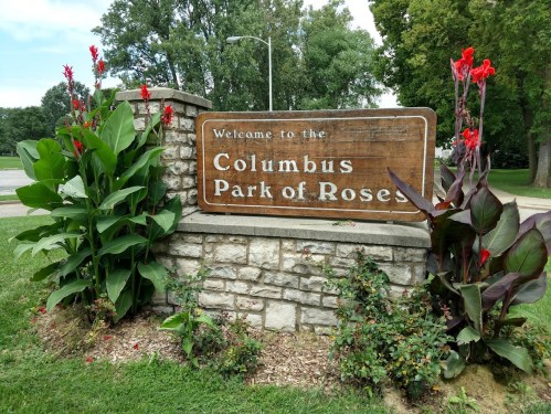 Sign welcoming visitors to Columbus Park of Roses, surrounded by vibrant red flowers and greenery.