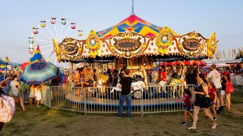 A vibrant carnival scene with a colorful carousel, Ferris wheel, and crowds enjoying the festivities under a clear sky.