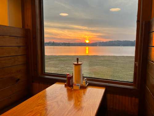 A cozy restaurant table by a window, overlooking a sunset reflecting on a calm lake.