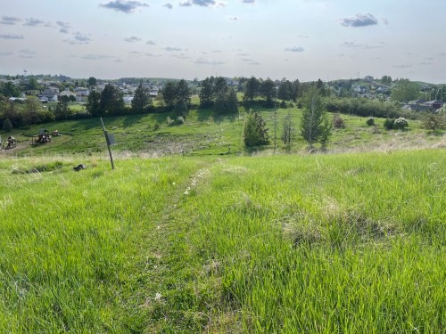A grassy path leads down a hill, with trees and a distant view of a small town under a clear sky.