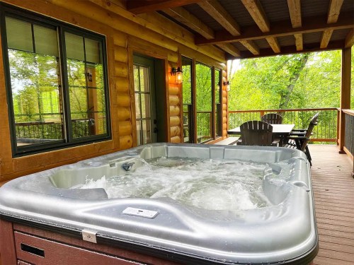 A hot tub on a wooden deck with a view of trees, surrounded by log cabin walls and outdoor seating.