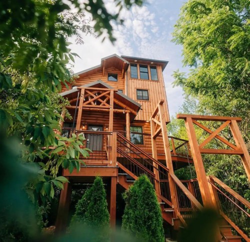 A tall wooden house surrounded by greenery, featuring a large deck and multiple windows under a blue sky.
