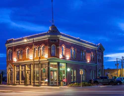 Historic brick building illuminated at dusk, featuring decorative lights and large windows, set on a street corner.