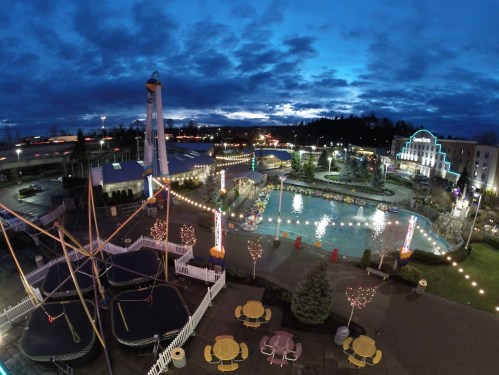 Aerial view of a vibrant amusement park at dusk, featuring a pool, rides, and colorful lights against a cloudy sky.