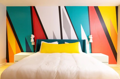 A modern bedroom featuring a colorful geometric accent wall and a white bed with yellow and white pillows.