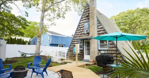 Charming shingle-sided A-frame cottage with a blue patio set, fire pit, and bright teal umbrella.