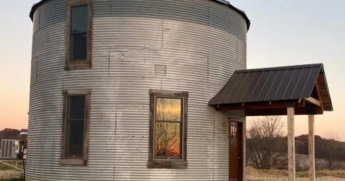 A tall, rustic metal grain silo transformed into a unique multi-story living space with a modern front door.