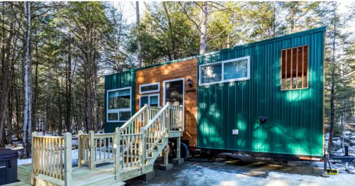 A modern tiny home with green metal siding and wood accents, parked in a snowy, wooded setting.