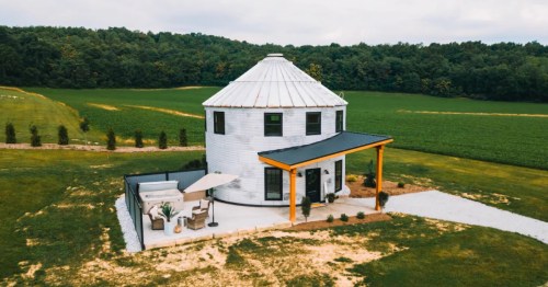 Unique white grain silo converted into a home with a patio and hot tub in a green field.