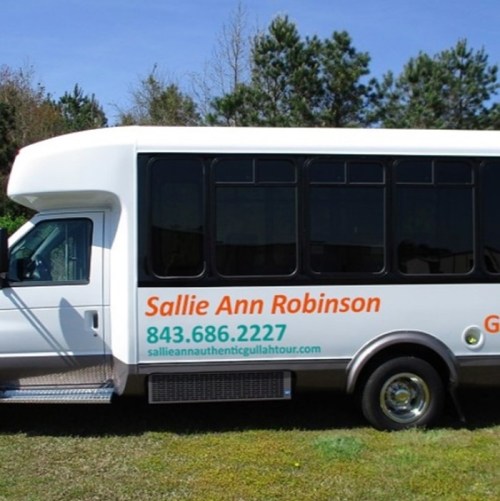 A white tour bus with green and orange text, parked in a wooded area. The bus advertises Sallie Ann Robinson's tours.